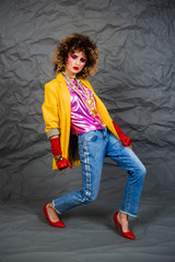 Girl in a yellow jacket and blue jeans with an afro hairstyle. Fashion of the eighties, disco era. Studio photo on gray background.