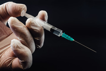 Close up of hand holding syringe. Doctor or nurse wearing rubber gloves and holding a syringe with attached needle. Covid-19 corona vaccine, medical healthcare and treatment concept, black background.