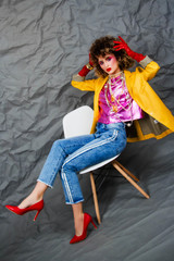 A girl in a yellow jacket and blue jeans with an afro hairstyle sits on a chair. Fashion eighties, the era of disco. Studio photo on a gray background.
