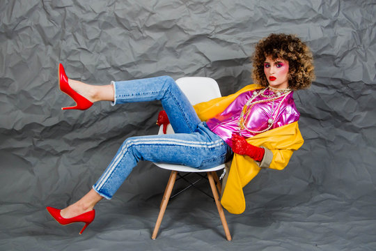 A girl in a yellow jacket and blue jeans with an afro hairstyle sits on a chair. Fashion eighties, the era of disco. Studio photo on a gray background.
