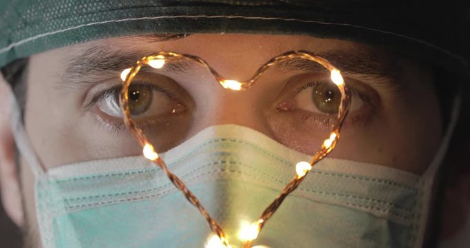 Primo piano macro degli occhi stanchi e sanguinanti di un medico che sta curando i pazienti affetti da Coronavirus in reparto di terapia intensiva. Tiene in mano un piccolo cuore luminoso.