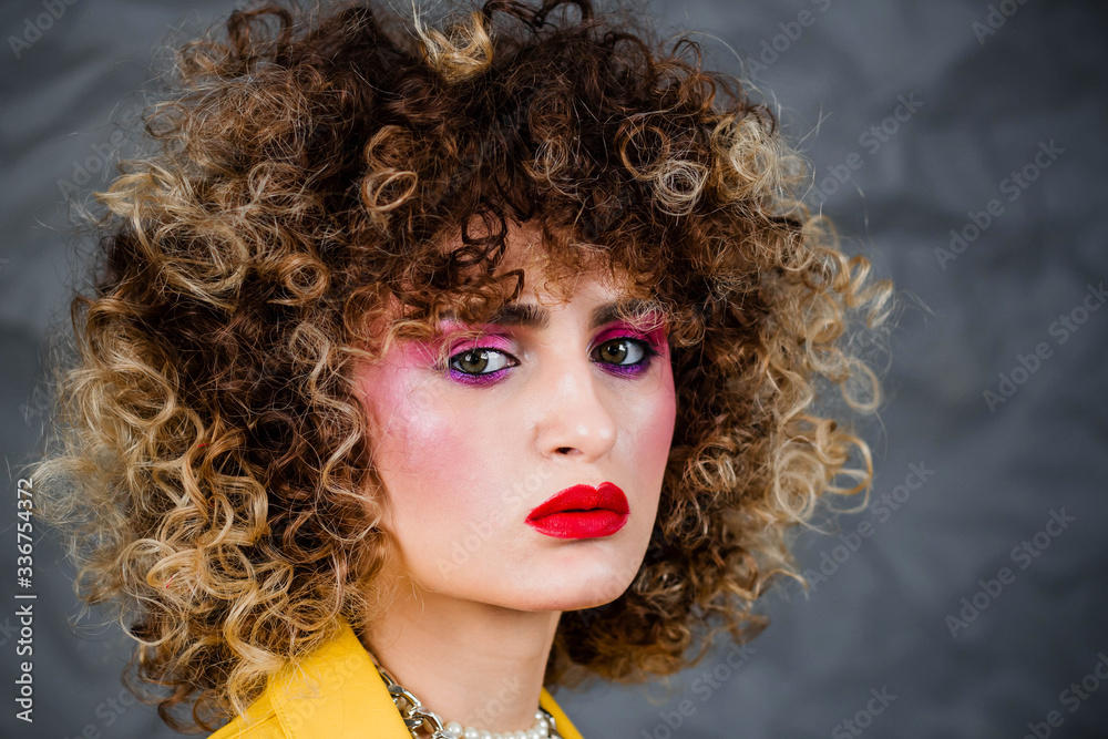 Wall mural Portrait of a girl in a yellow jacket and blue jeans with afro hair of the eighties, disco era. Photo in studio on a gray background.