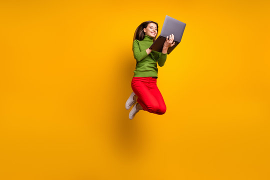 Full Length Photo Of Funny Pretty Lady Jumping High Holding Using Notebook Modern Technology Wear Casual Green Jumper Red Pants Isolated Yellow Color Background