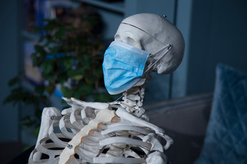 The human skeleton in a medical mask sits on a chair at home. The concept of a coronavirus pandemic.