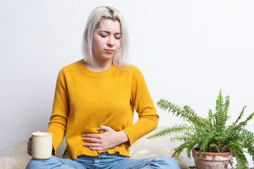 Young blonde female sitting at home holding a cup of tea while putting her hand in her stomach