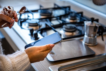Mani di ragazza che controllano un o smartphone mentre in cucina si prepara il caffè nello moka sullo sfondo