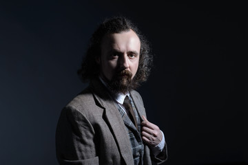 studio dramatic of a young bearded and curly-haired guy of thirty years, in an expensive business suit, holds his hand by the lapel of his jacket. On a dark background.
