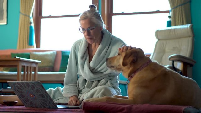 Mature woman on the floor in bathrobe talking to and petting dog then typing on laptop computer while on the floor.
