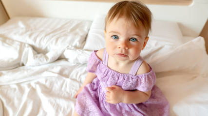 Close up portrait of sad blue-eyed toddler. Baby looks to the camera sitting on the bed. Girl in dress looking up.