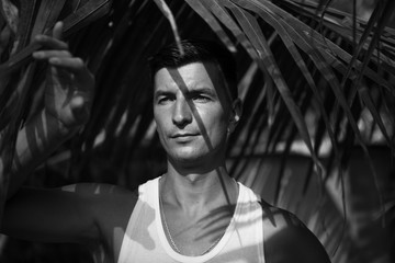 A young man with dark hair under a palm leaf with chiaroscuro from the branches on his face. A man in a tropical garden in India-a close-up portrait.