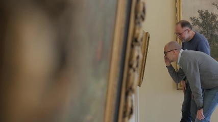 Two men in an art gallery look at a painting and talk about art