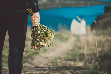 in the foreground in focus, the bride holding a bouquet of flowers, in defocus blur the bride hurries to the groom along the path. Against the background of the natural landscape.