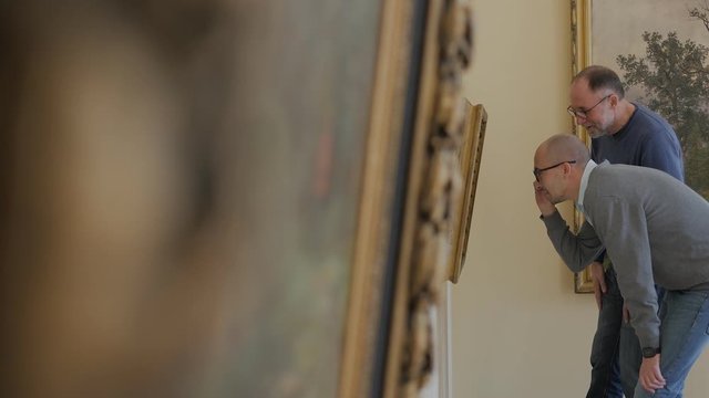 Two men in an art gallery look at a painting and talk about art