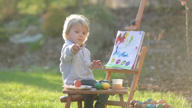 Sweet toddler child, painting eggs in garden with little chicks running around him on sunset
