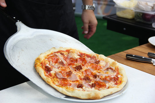 Italian cuisine. Pizza with hands of chef from the oven on the spatula. Dough, ketchup, bacon, salami, sausage and cheese on a white board on a wooden table. Background image, copy space