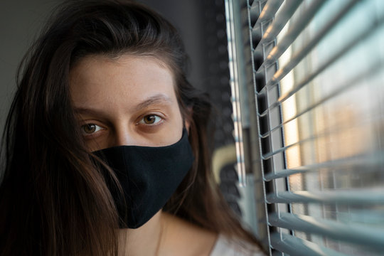A girl in a black medical mask sits on a windowsill and looks into the camera. Sunset light. Self-isolation