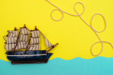 Toy ship is floating on the blue paper-cut waves, rope is twisted by loops above a ship on the yellow background.Concept summer and travel flat lay, copy space for mock-up