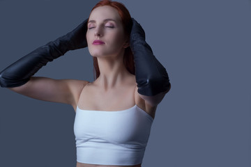 Naklejka premium Portrait of a young beautiful red-haired girl of thirty-five years old, in black leather gloves and a white tank top, touching her head with her hands, the model poses for a catalog magazine.