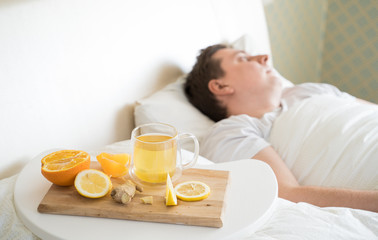 Fototapeta na wymiar Cup with antipyretic drugs for colds,flu.Sick man in bed. Tea with citrus vitamin C,ginger root,lemon,orange.Wooden tray. Home self-treatment.Medical quarantine antiviral covid-19 coronavirus therapy