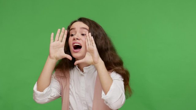 Attention, important advertisement! Cute teenage brunette girl in white shirt holding hands near wide open mouth and shouting loudly, screaming announcement. studio shot isolated on green background