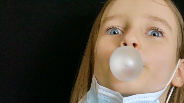 Coronavirus Covid-19 outbreak. Little girl is looking at the camera with happiness in the eyes and bubble gum in the mouth. Stay positive concept during self-isolation at home in protective mask