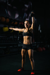 Fitness young woman with perfect muscular body in black sportswear is lifting kettlebell overhead during weight training workout. Concept of healthy lifestyle and workouts in a modern dark gym.