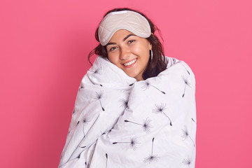 Image of brunette smiling young girl posing wrapped in white blanket, having blindfold on her forehead, looking directly at camera, being happy stay at home, resting in bed, female looks satisfied.
