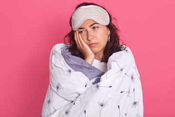 Tired woman standing with palm on cheek, looking aside with exhausted facial expression, posing wrapped in plaid and wearing blindfold, being bored, stands isolated over pink studio background.