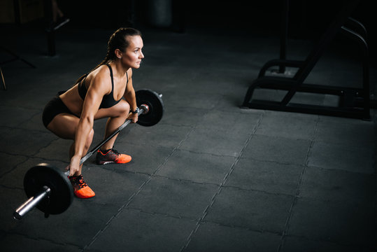 Athletic woman bodybuilder with perfect fitness body preparing to lift the heavy barbell from the floor. Concept of healthy lifestyle and workouts in a modern dark gym.