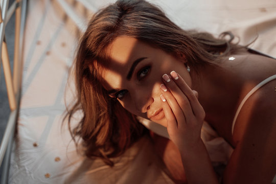 Fit and sexy woman with brown curly hair laying among bed sheets with star-shaped confetti, wearing white ingerie and looking hot. Close up look