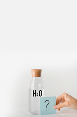 Glass bottle with the inscription chemical formula of water on it and question mark on a note sheet.