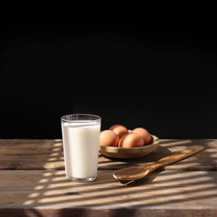 brown eggs lie on a wooden bowl, an old spoon and a glass of white milk in the rays of light on the table, retro concept