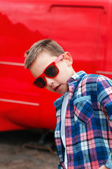 Portrait little boy wearing a checkered shirt and sunglasses in city over red background