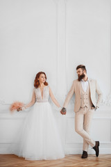 a guy and a girl in a white dress stand against a white wall and hold hands
