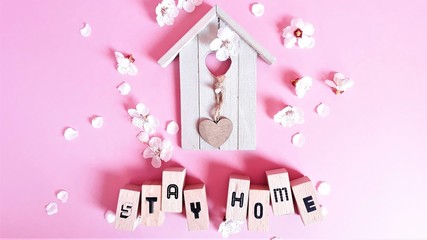 Wooden decorative house and wooden letters with text stay home,stey safe among apricot flowers on a pink background.View from above.