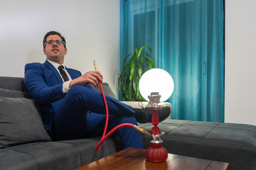 Young man sitting on a couch with his legs crossed. He smoking with a shisha. He is wearing a blue suit. The hookah is red.