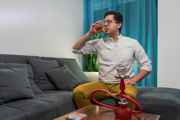 Young man sitting on a sofa. He smoking a shisha and drinking whiskey.