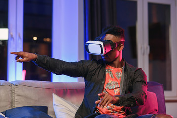 Excited young african-american man extending hand forward, using virtual reality headset sitting on bed, copy space
