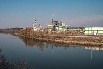 Fototapeta premium pipes of woodworking enterprise plant sawmill near river. Air pollution concept. Industrial landscape environmental pollution waste of thermal power plant