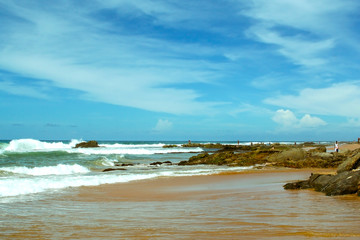 Praia de Salvador em dia bonito de sol com banhistas distantes sobre as pedras enquanto ondas quebram nas rochas.