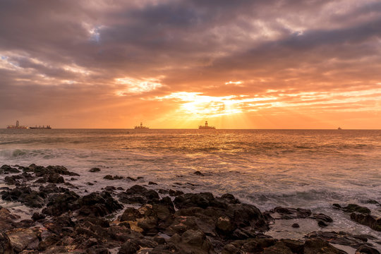 Sunrise from a stony shore to boats moored on the horizon with the rising sun and pesky shining through the clouds to the ocean with moving clouds from the Grand Canary Island.