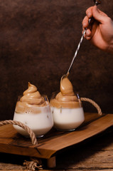 Dalgona cold whipped coffee with milk foam, on wooden tray with hand, instant coffee, Korean trend. Home-made recipe in the process. On wood table, brown colors, with place for text and other design.