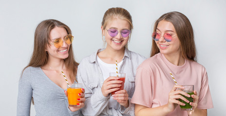 Three cheerful girls drinking fresh raw detox juice on gray