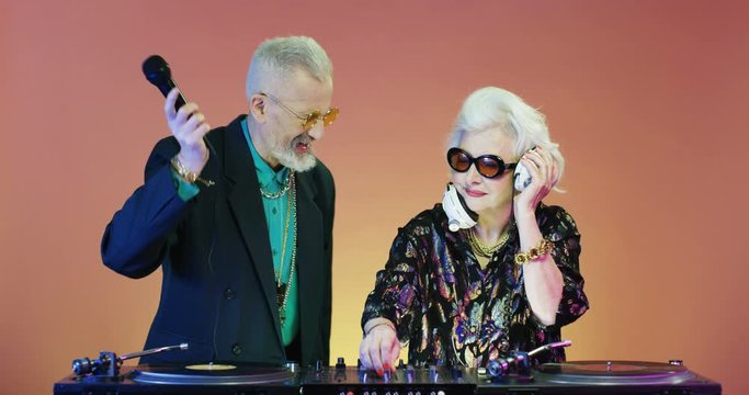 Glamorous vintage stylish grandmother playing music at DJ mixer on pink wall background and grandfather singing in mic. Couple of old retro male and female djs playin music on equipment.