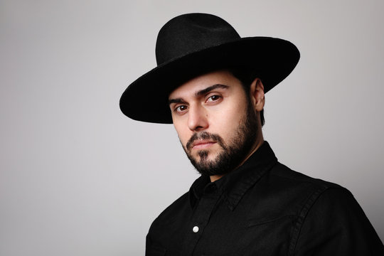 Side portrait of bearded hipster wearing black fedora hat and shirt looking at camera.