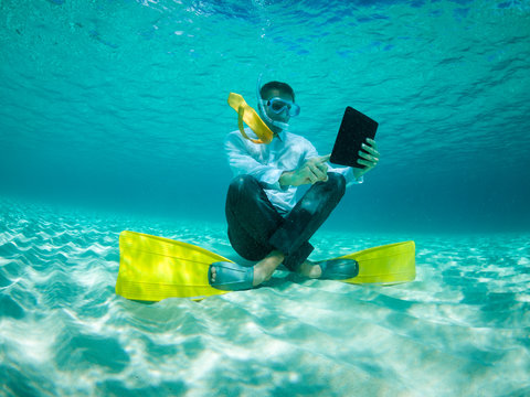 Snorkelling businessman in shirt and tie and matching fins using a tablet computer sitting underwater in tropical turquoise sea