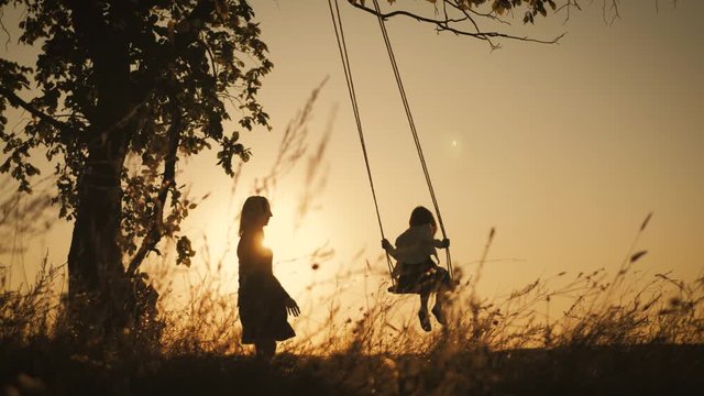 Silhouette of happy young mother and little daughter on a swing at sun light. Pretty girl sitting on a wooden swing and looking at golden sunset. Happy family concept.