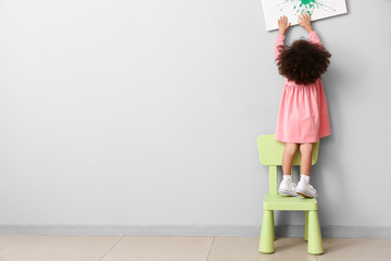 Little African-American girl standing on chair near light wall. Child in danger