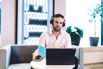 Skilled Caucasian man watching video via portable touch pad connecting to 4g wireless internet, male in electronic headphones for noise cancellation reading publication text on digital tablet