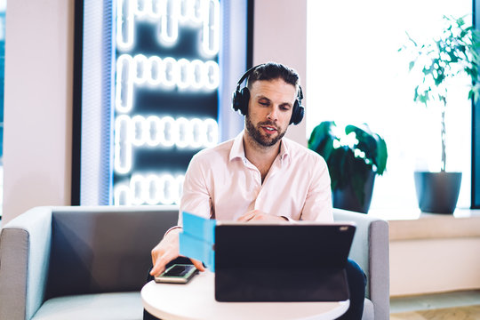 Skilled Caucasian man watching video via portable touch pad connecting to 4g wireless internet, male in electronic headphones for noise cancellation reading publication text on digital tablet